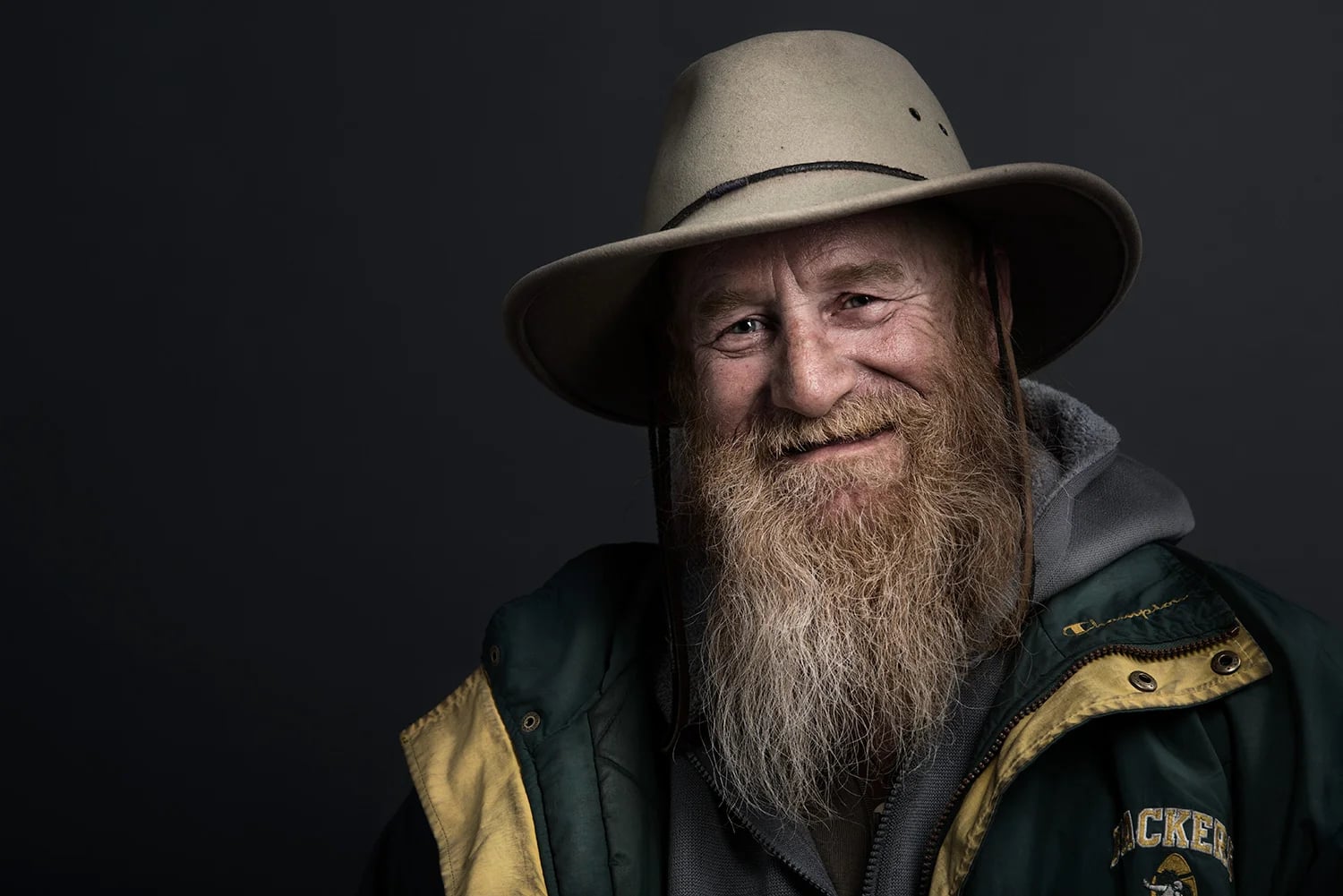 A bearded man wearing a wide-brimmed hat, a gray hoodie, and a green jacket with yellow lining smiles warmly against a dark background.