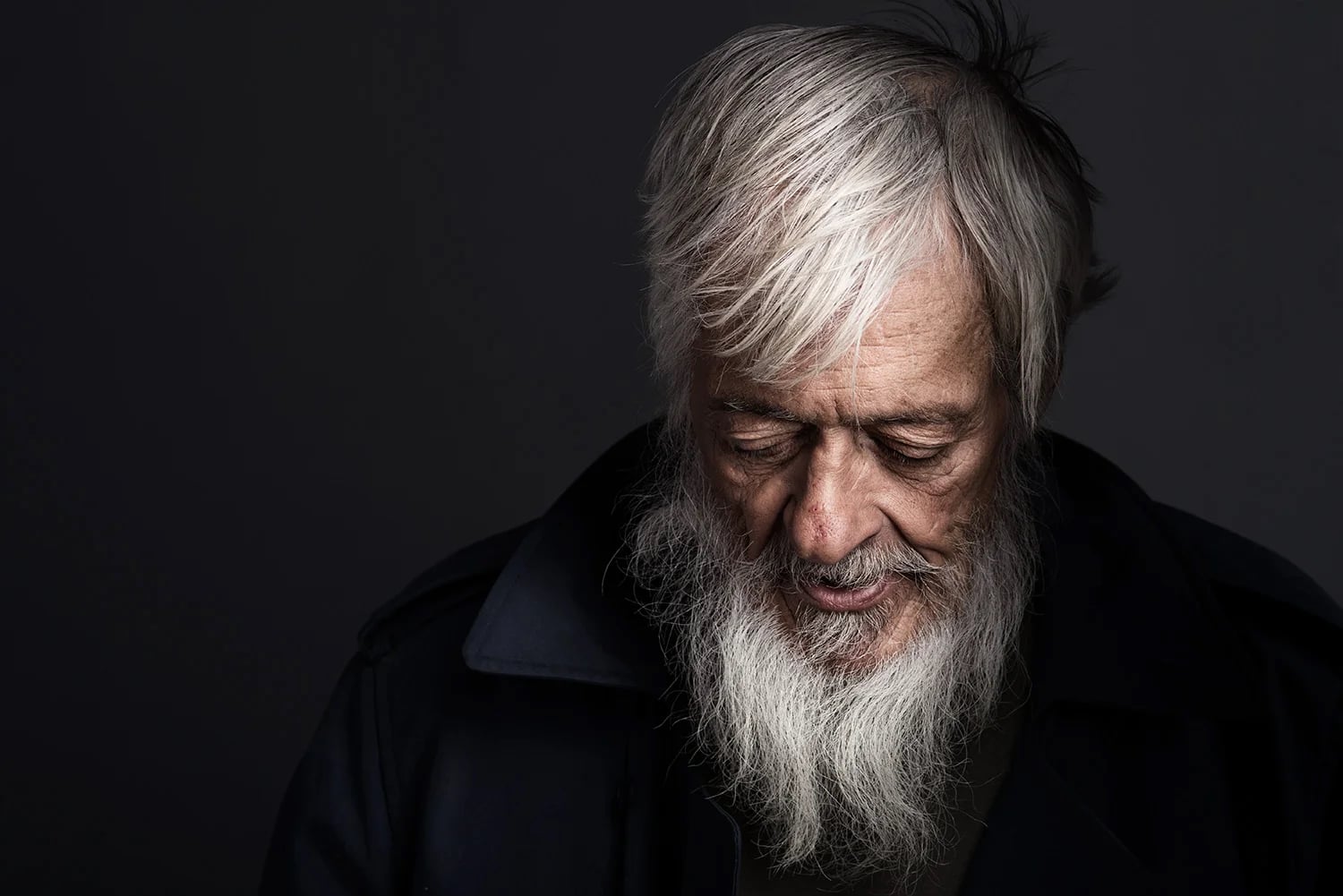 An elderly man with white hair and a long white beard looks down with a gentle expression. He is wearing a dark coat, and the background is dark, highlighting his thoughtful demeanor.