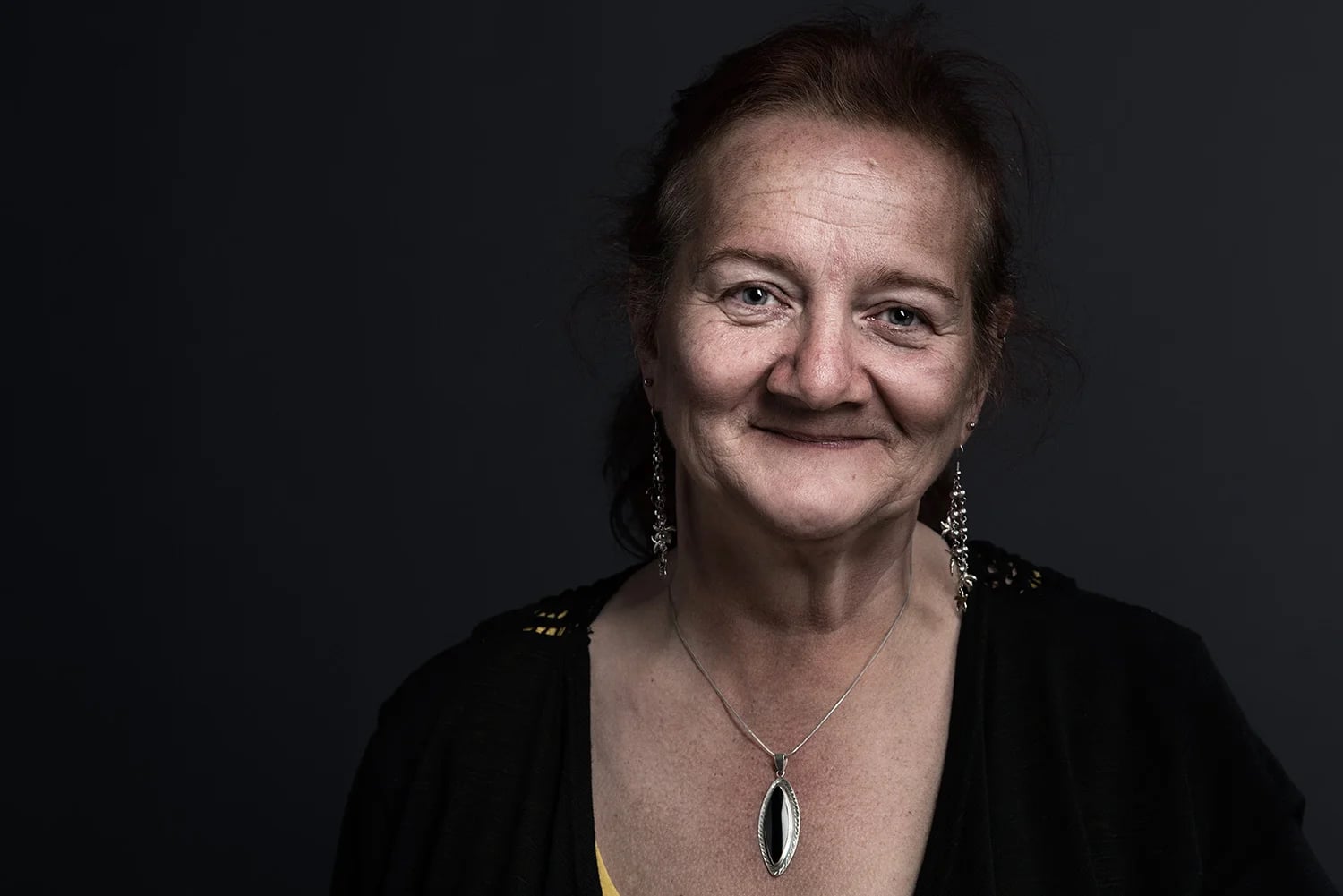 A middle-aged woman with light skin and reddish-brown hair smiles softly against a dark background. She wears dangling earrings, a necklace with a stone pendant, and a black top.