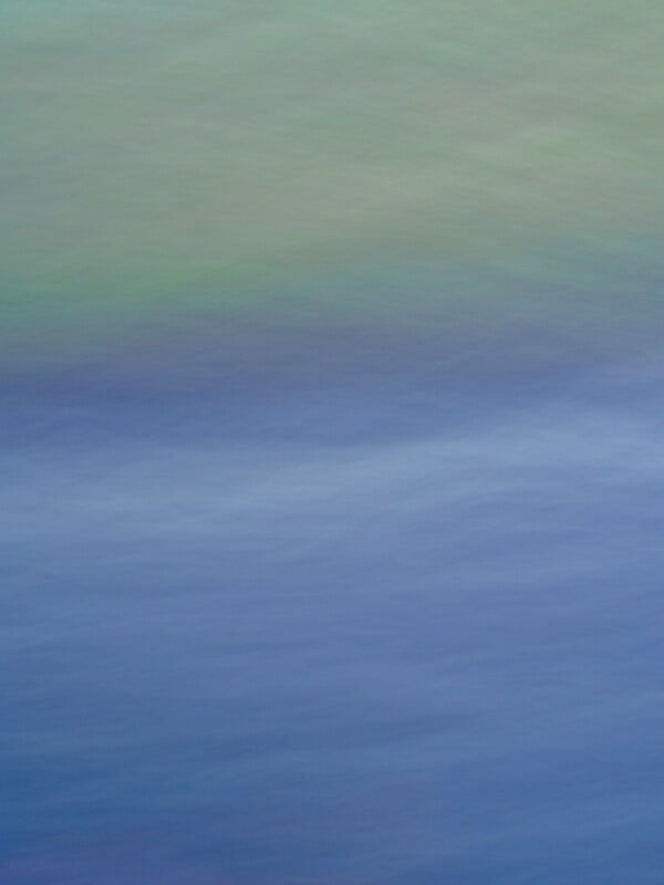 A soft, abstract image featuring blurred horizontal bands of blue, purple, and green, resembling gentle waves or a serene landscape with water and sky.