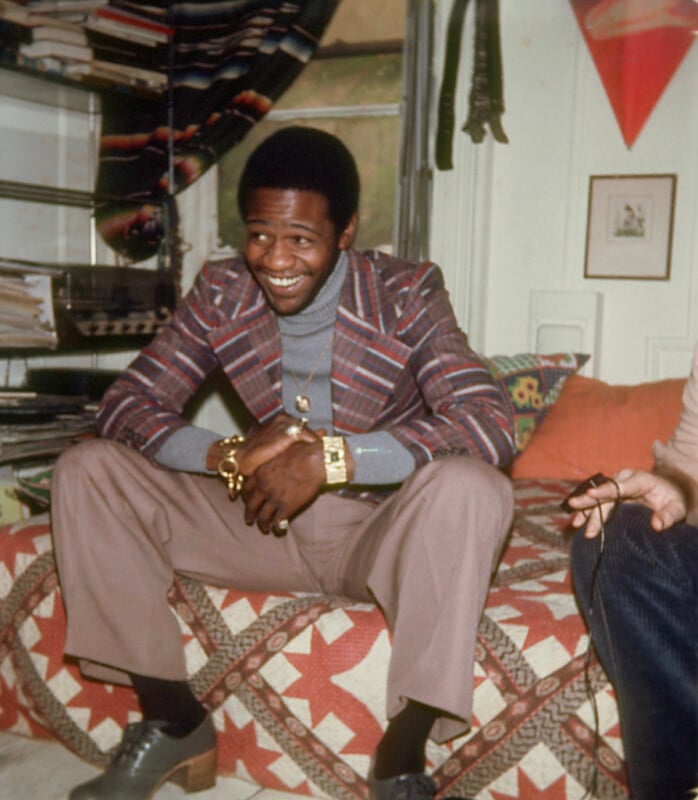 A man wearing a plaid blazer, gray turtleneck, and brown pants sits smiling on a quilt-covered bed in a cozy room with shelves, books, and wall art visible in the background.