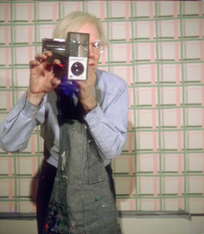 A person with light hair, wearing glasses, a blue shirt, and a paint-splattered apron holds a vintage camera up to their face. The background features a patterned wall with a geometric pink and green design.