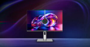 A sleek ASUS monitor displays vibrant abstract digital art, with a round webcam mounted at the center top. The monitor stands on a modern silver base against a dark, colorful, gradient background.