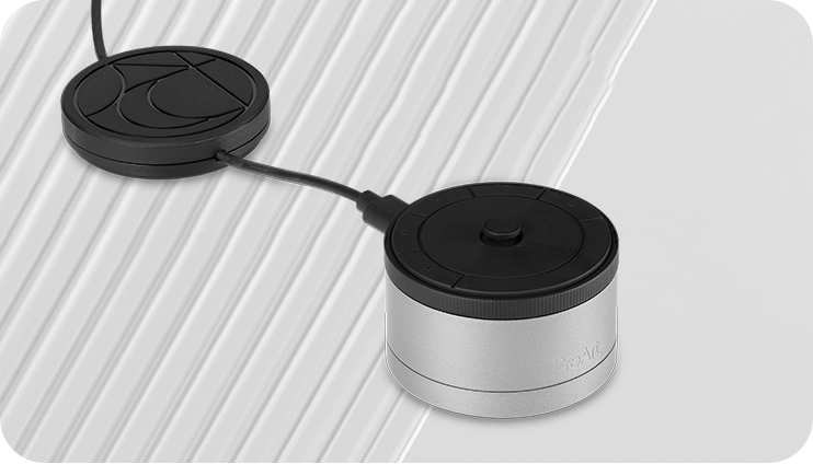 Un dispositivo electrónico redondo, plateado y negro conectado mediante cables a una almohadilla plana y redonda de color negro con un diseño geométrico, colocada sobre una superficie blanca inclinada y estriada.