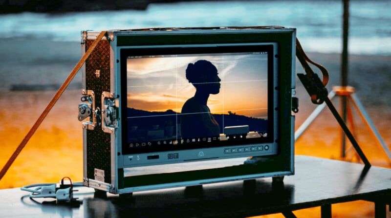 A monitor on a table displays the silhouette of a woman against a sunset, with a beach and ocean visible in the background. The scene is framed as if being filmed or photographed.