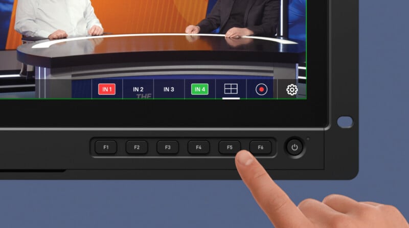 A person’s finger presses the “F6” button on a monitor below a live video feed showing two people at a news desk. The screen displays camera input buttons labeled IN 1, IN 3, and IN 4.