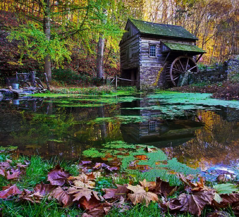 A rustic wooden mill with a water wheel stands beside a pond covered in green moss. Autumn leaves are scattered on the grass in the foreground, and trees with fall foliage surround the scene, reflected in the still water.