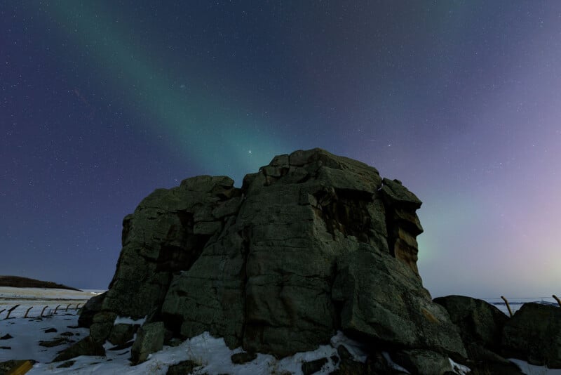 Large rocky outcrop on a snowy landscape under a starry night sky, with faint green aurora borealis lights stretching overhead.