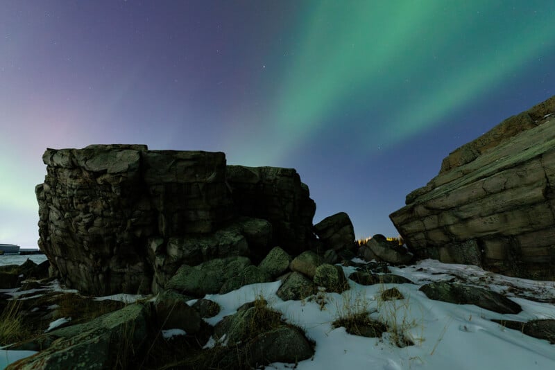 Rocky terrain with patches of snow under a night sky illuminated by green northern lights, with large boulders and cliffs in the foreground and faint light on the horizon.