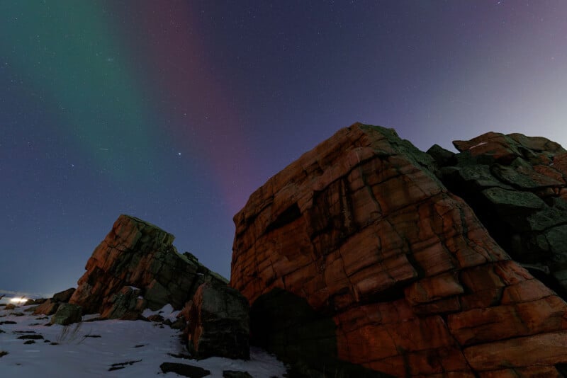 Large reddish rock formations stand beneath a night sky with faint green aurora and scattered stars; patches of snow are visible on the ground.
