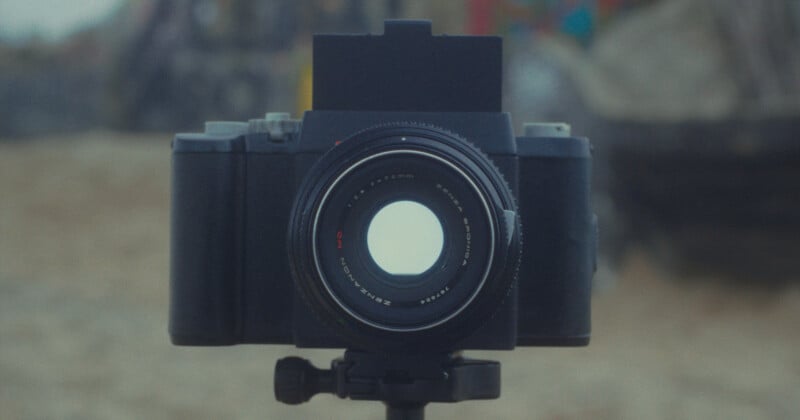 A close-up view of a black camera mounted on a tripod, with the lens facing forward. The background is blurred, highlighting the camera as the main subject.