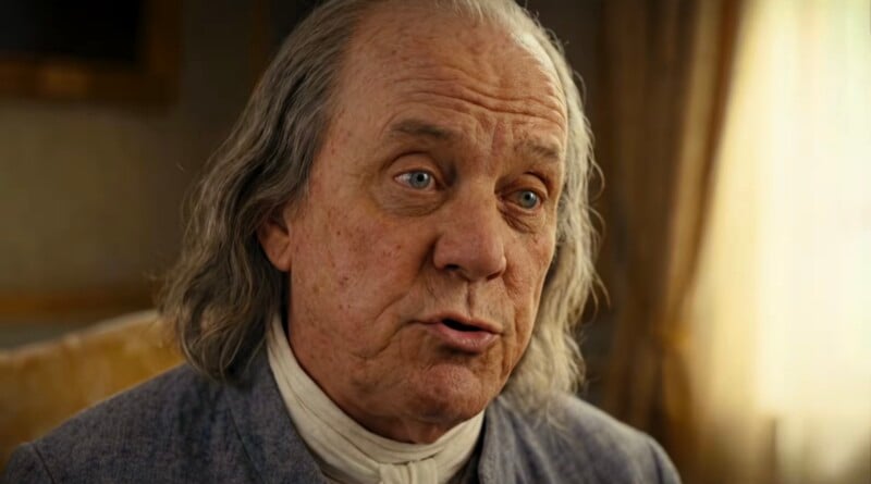 An older man with long gray hair and a wrinkled face, dressed in 18th-century clothing, sits indoors with soft light coming through a window behind him.