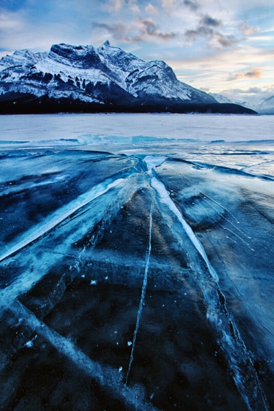 Cracked blue ice covers a frozen lake with dramatic ice lines, set against snow-covered mountains and a cloudy sky at sunset.