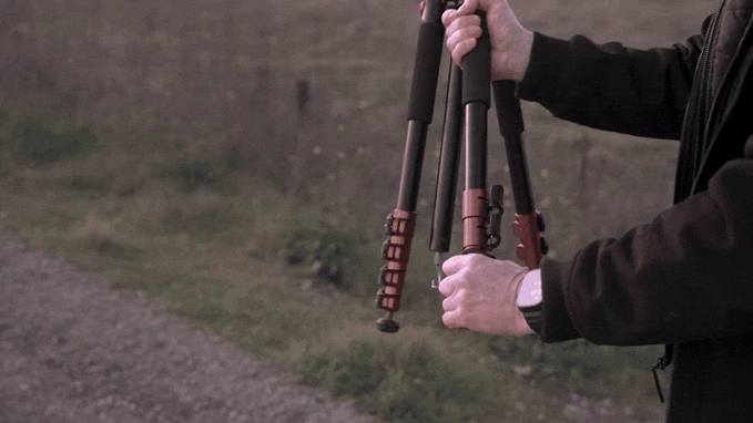 A person outdoors rapidly sets up a compact tripod, extending and locking its legs with swift movements. The background is a grassy field.
