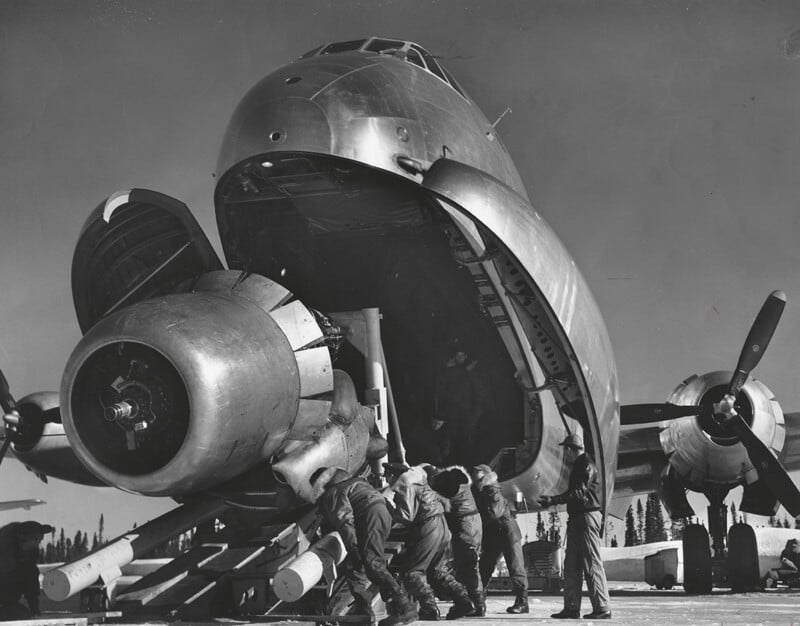 A group of soldiers loads supplies into the open nose cargo hold of a large military transport aircraft, with its propeller engines and wheels visible, on an outdoor airfield.