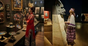 Two women in traditional clothing admire artifacts in a museum; one wears a red embroidered gown and studies ornate plates and paintings, while the other in a purple outfit observes a large stone sculpture.