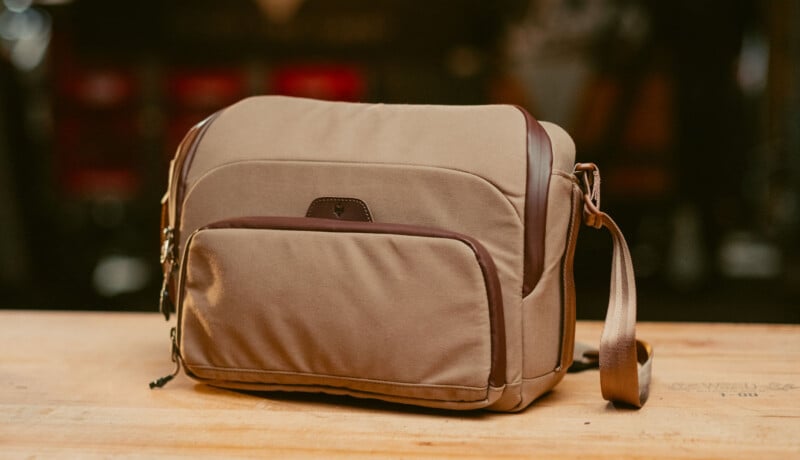 A tan and brown shoulder bag with multiple zippered compartments sits on a wooden surface, with a blurred background.
