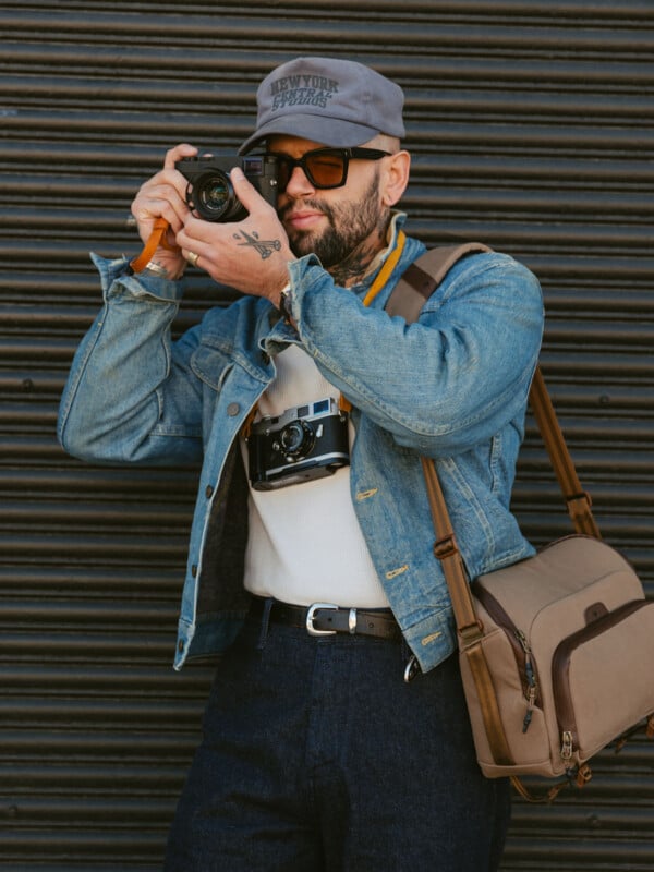 A man wearing sunglasses, a denim jacket, and a gray cap holds a camera up to his face. He has tattoos, a brown shoulder bag, and another camera hanging around his neck, standing in front of a black metal shutter.