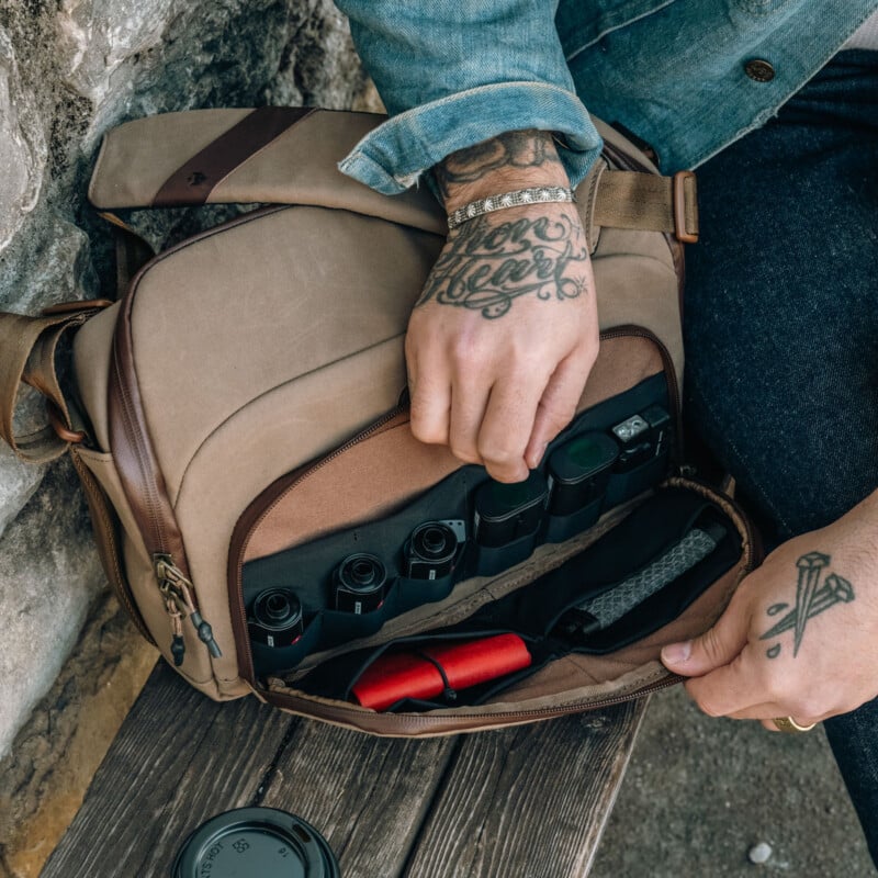 A person with tattooed hands opens a brown camera bag containing several camera lenses and accessories. The person is wearing a denim jacket and dark pants, sitting outdoors on a stone surface.
