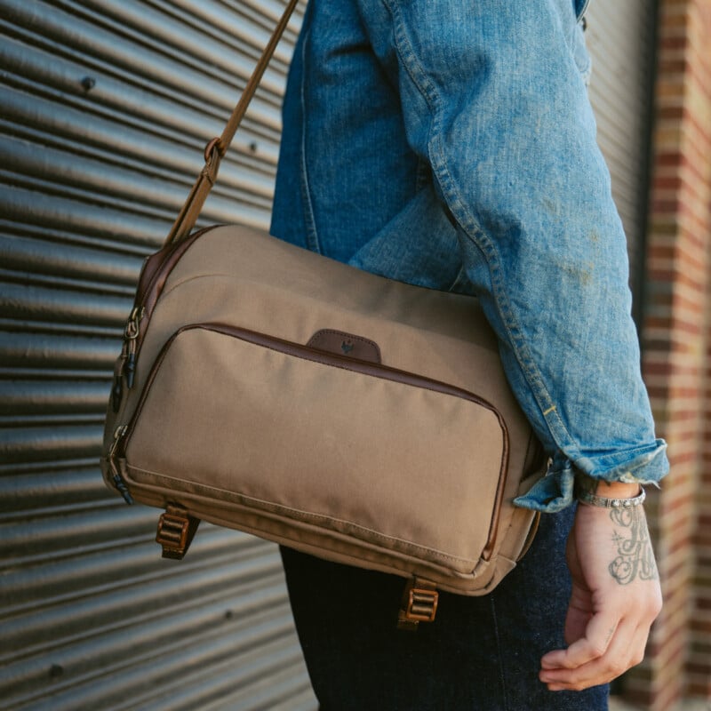 A person wearing a blue denim shirt and dark pants carries a brown messenger bag with leather accents over their shoulder, standing next to a ridged metal wall. A tattoo is visible on their hand.