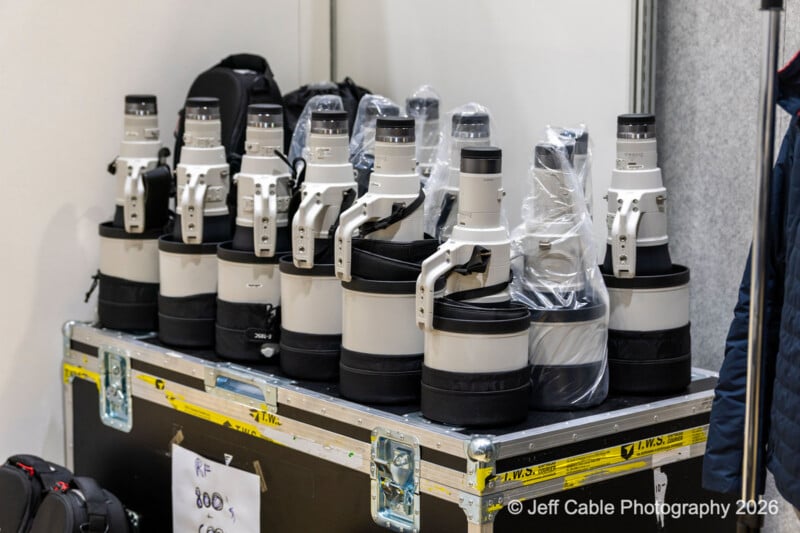 A row of large white telephoto camera lenses, some covered in plastic, arranged on a hard case with backpacks nearby. A copyright watermark reads "© Jeff Cable Photography 2026".