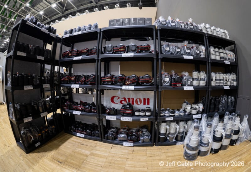 Large black shelves filled with Canon cameras, camera bodies, and various camera lenses, many labeled and some wrapped in plastic, displayed in an indoor setting with a "Canon" banner in the center.