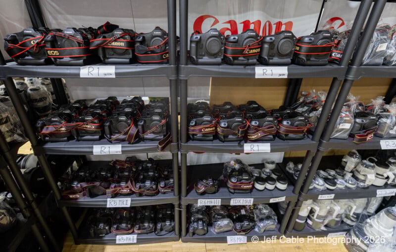 Several rows of black Canon cameras with red straps and various lenses are organized on dark shelves, each labeled with white tags. Some camera equipment is in plastic bags on the right side.