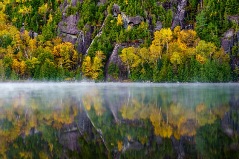 A calm lake reflects trees with green and yellow autumn foliage, as well as rocky cliffs, under a misty layer at the water’s surface.