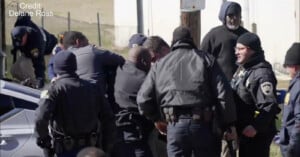 A group of police officers gather closely around a person near a car on a street during daytime. Some officers appear to be restraining the individual while others observe the scene. Credit: Delane Ross.
