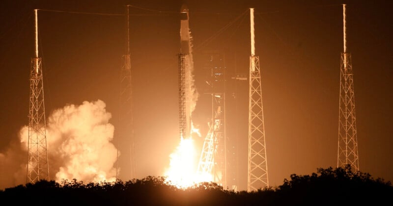 A rocket launches into the night sky, surrounded by tall support towers. Bright flames and smoke billow from the base, illuminating the scene against the dark backdrop.
