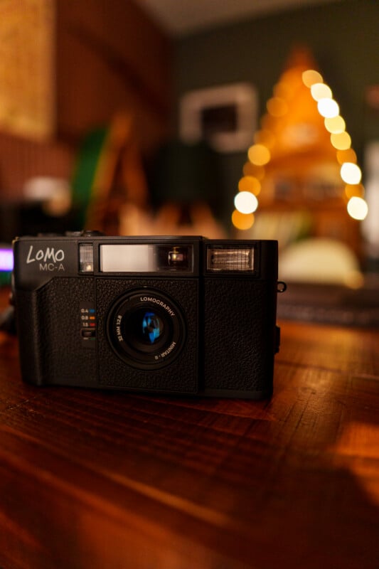 A black Lomo camera sits on a wooden table in a warmly lit room, with blurred yellow fairy lights and a triangular wooden structure in the background.