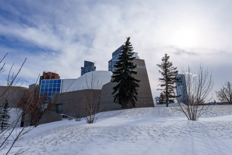 A modern building with geometric shapes stands behind snow-covered ground and a few tall pine trees, with city skyscrapers visible in the background under a partly cloudy sky.