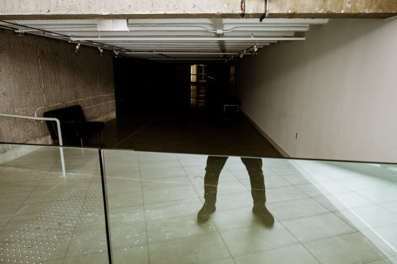 A glass barrier reflects a person's legs and feet in an empty, dimly lit hallway with concrete walls and minimal furniture.