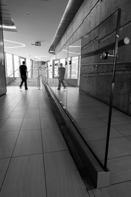 Black and white photo of a modern hallway with tiled floors and concrete walls. A person walks away from the camera, and their reflection appears in a glass railing on the right side. Light streams in from windows ahead.