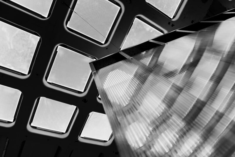 Black and white photo of a geometric ceiling with square light panels and an angled, reflective glass or plastic structure in the foreground, creating abstract, modern architectural patterns.