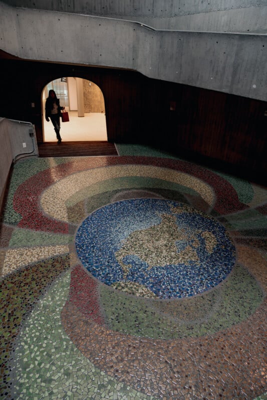 A person walks through an arched doorway above a mosaic floor depicting a map of the world in blue, green, and brown tiles, set within a circular design in a concrete-walled building.
