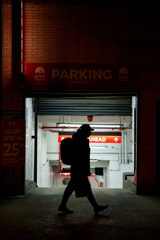 A person with a backpack is silhouetted while walking past the entrance to an indoor parking garage at night, illuminated by bright red and white signs.