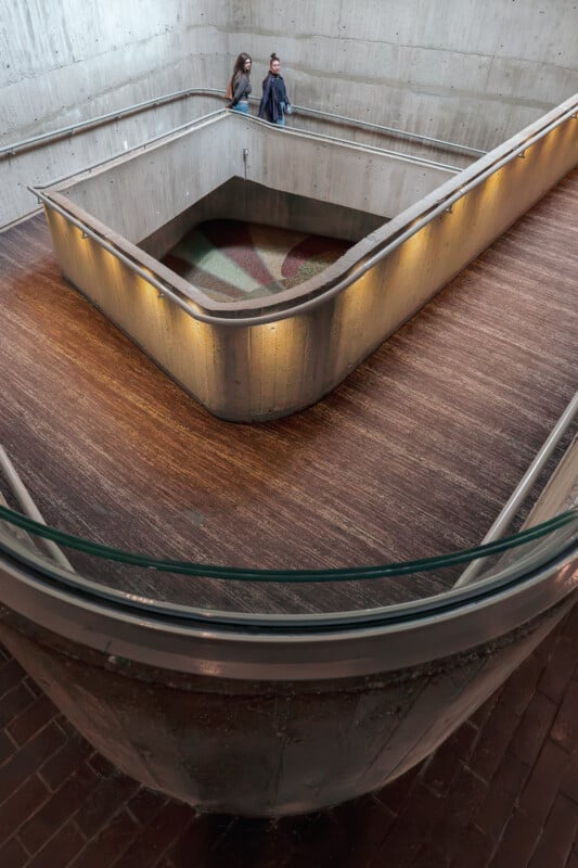 Two people stand on a concrete ramp inside a modern, geometric, multi-level indoor space with textured flooring and curved railings. The area is lit by wall lights and features minimalistic, industrial design.