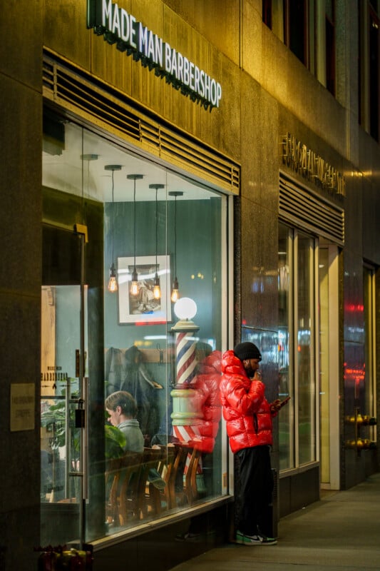 A person in a bright red puffer jacket stands outside Made Man Barbershop at night, looking at their phone. The shop’s lit interior and a traditional barber pole are visible through the window.