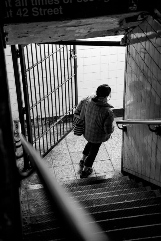 A person wearing a jacket and carrying bags descends a tiled staircase into a subway station. The image is black and white, and a metal gate and sign are visible in the background.