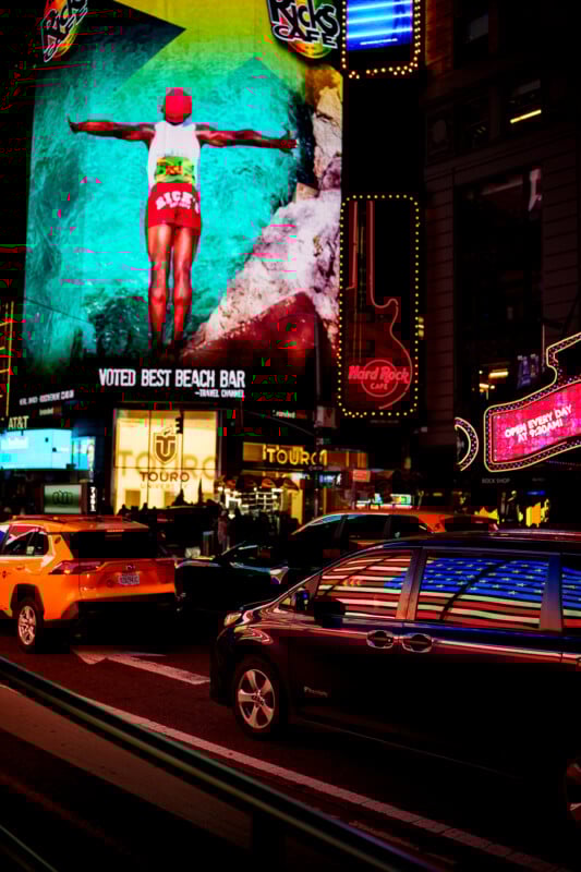 A vibrant city street at night with colorful neon signs and billboards, including a large ad of a person diving into a pool. Cars, including a yellow taxi, are stopped in traffic, reflecting neon lights.