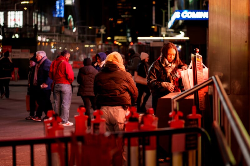A woman uses a ticket booth on a city street at night, illuminated by warm lights. Several people are gathered nearby, some wearing coats and hats, with city buildings and signs glowing in the background.