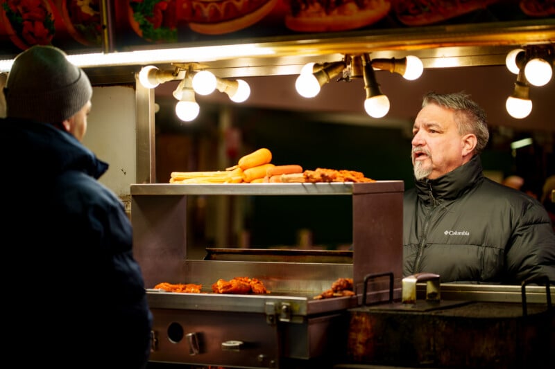 A vendor in a black jacket stands behind a food stall counter, talking to a customer in a beanie. Warm lights illuminate food items, including carrots, displayed on the counter.