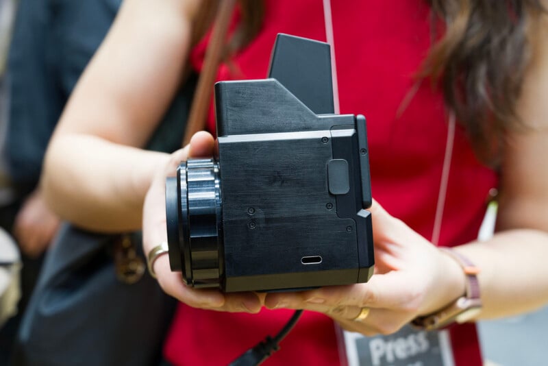 A person in a red shirt holds a modern instant film camera close to the viewer, showing its side and lens. The person's press badge is partially visible, and their face is out of frame.