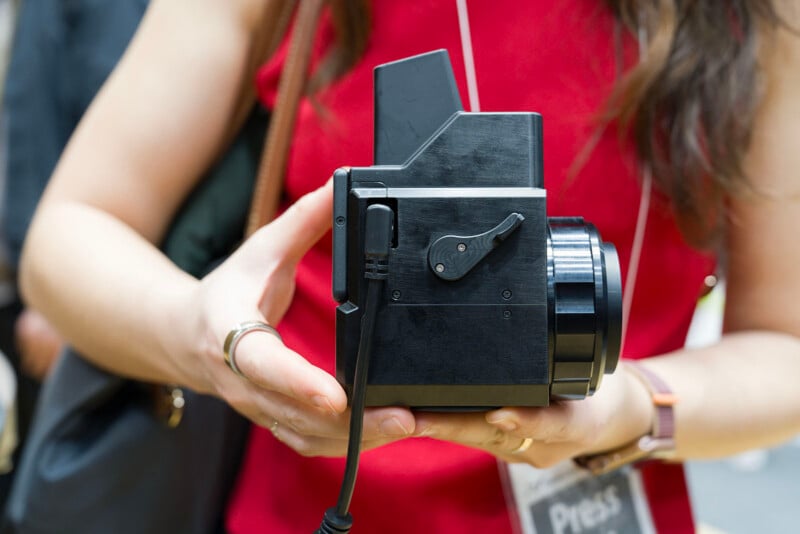 A person wearing a red top holds a black vintage instant camera with both hands. The camera has a large lens, a cable, and a lever on the side. The person's face is not visible.