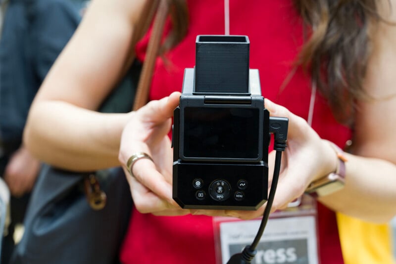 A person in a red top holds a black, boxy camera or electronic device with a small screen and buttons, displaying it towards the viewer. The person is wearing a watch and a badge.