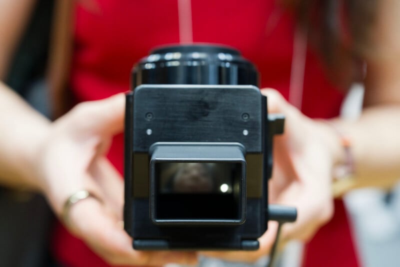 A person in a red shirt holds a black camera facing the viewer, with both hands visible. The camera lens and viewfinder are in focus, while the background and person's face are blurred.