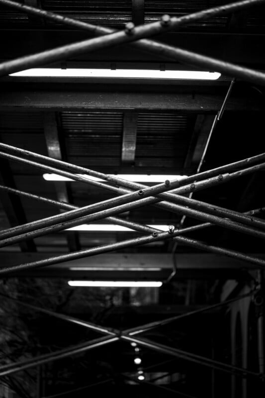 Black and white photo of scaffolding with intersecting metal pipes forming X shapes, illuminated by fluorescent lights from above, creating a geometric and industrial pattern.