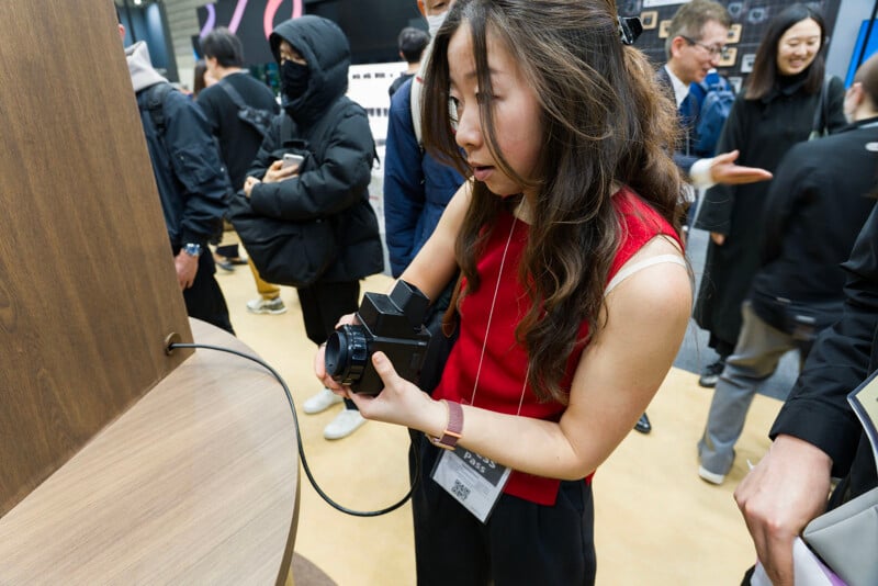 A woman in a red top examines a camera at an event, with a crowd of people around her. She appears focused, holding the camera with both hands near a wooden display.