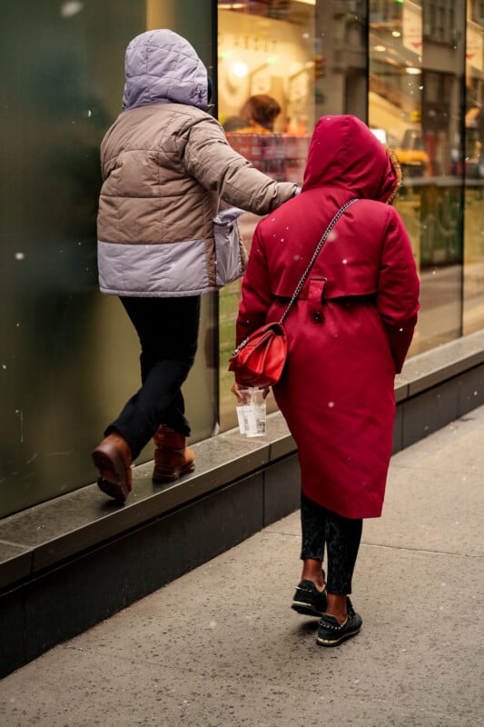 Two people in winter coats, one in a red coat and the other in a light purple coat, walk along a city sidewalk. The person in purple steps onto a narrow ledge beside a glass storefront.